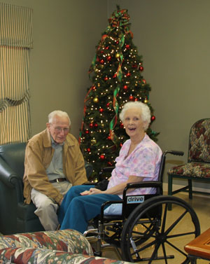 elderly couple sitting in front of a christmas tree smiling at the camera